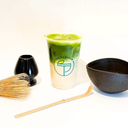 Uji matcha latte in a Tea Parlour cup, styled with traditional tea tools including bamboo whisk, scoop, chasen stand, and a black tea bowl on a white background.